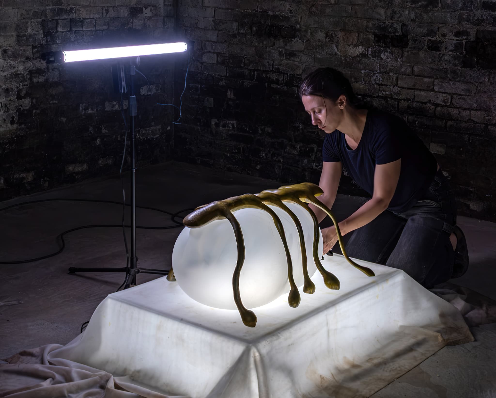 In a dark room, a woman kneels beside a sculpture of a bug illuminated from above and below.