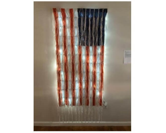 A vertical American flag displayed on a wall with lights shining through from behind.