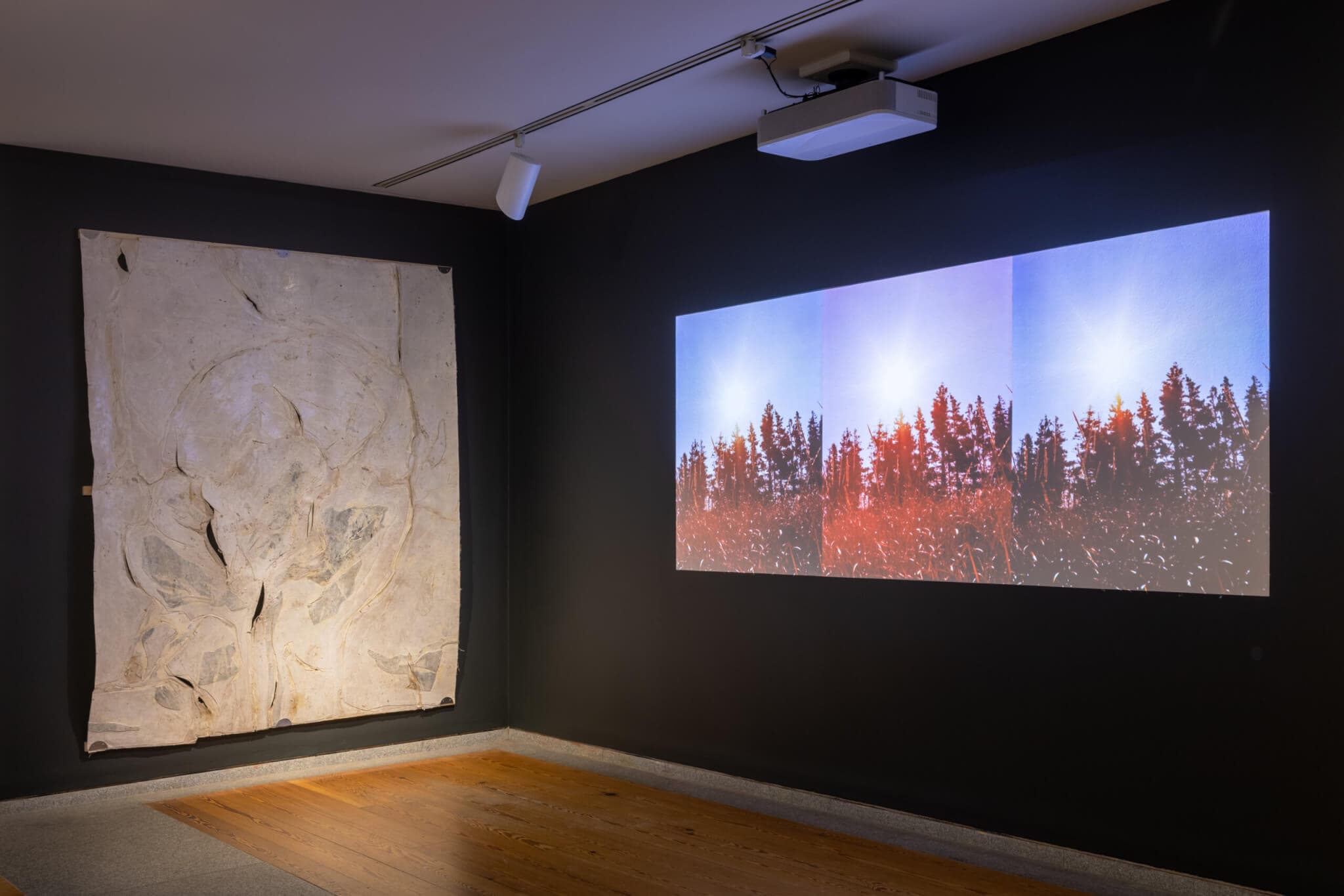 A video projection and wall installation are display on adjacent walls in a gallery space.