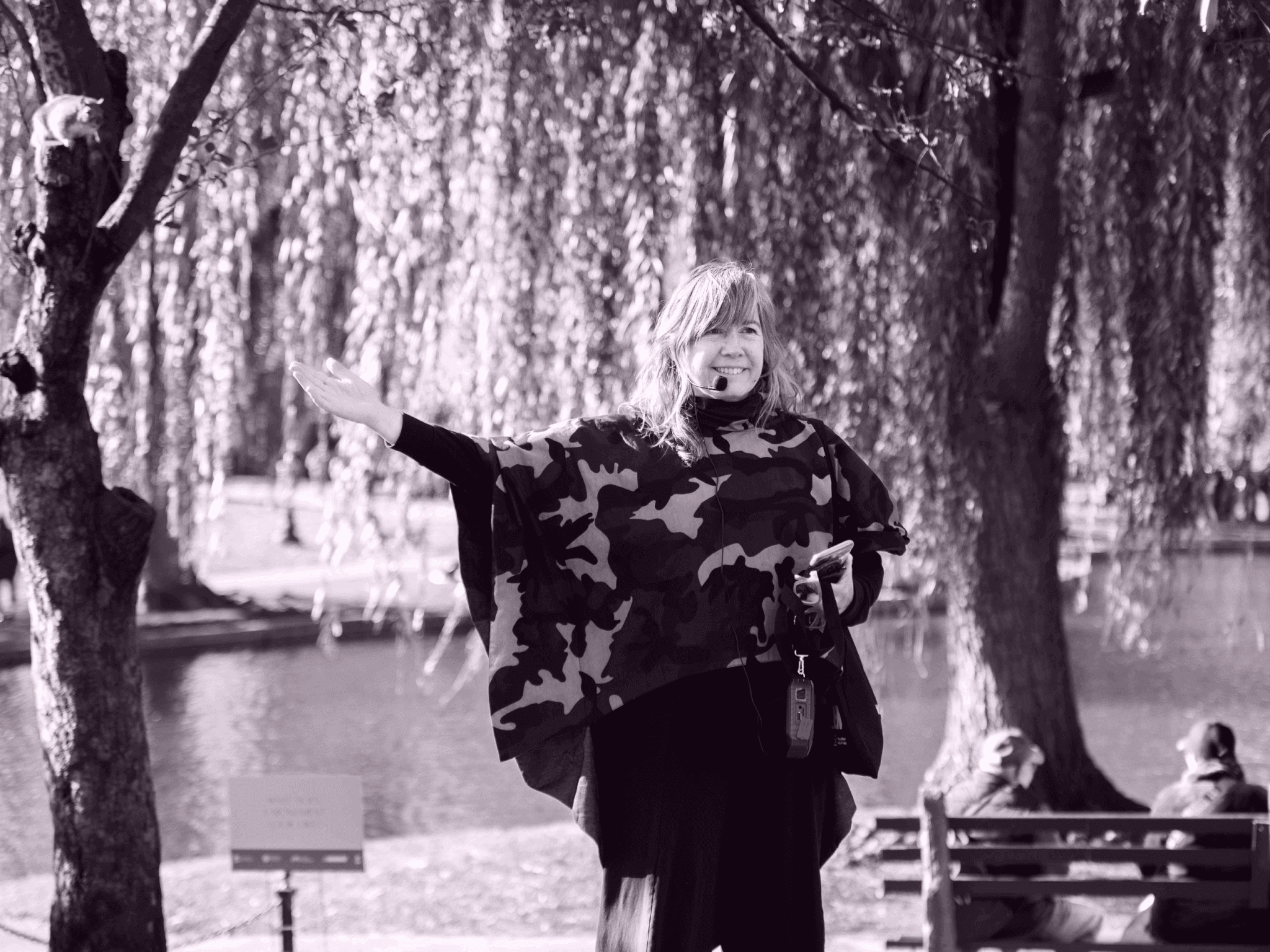 A woman with a microphone headset gestures toward the trees in a public garden.