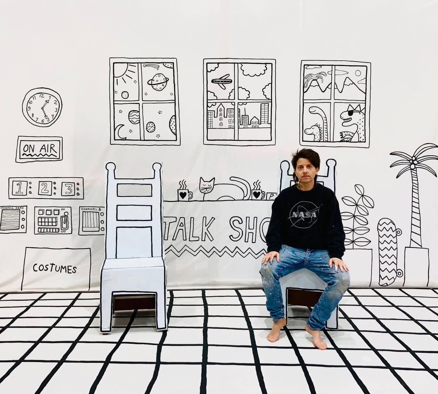 An artist wearing jeans and a sweatshirt sits in a hand drawn talk show set.